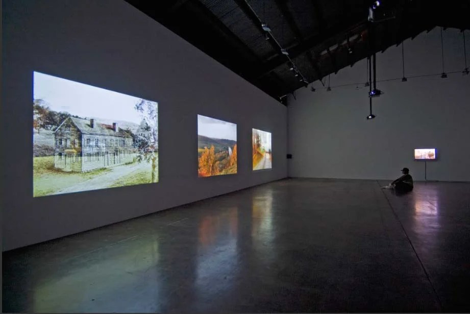 An art gallery with three large digital screens displaying landscape images and a person sitting on the floor watching a small television screen in the corner.
