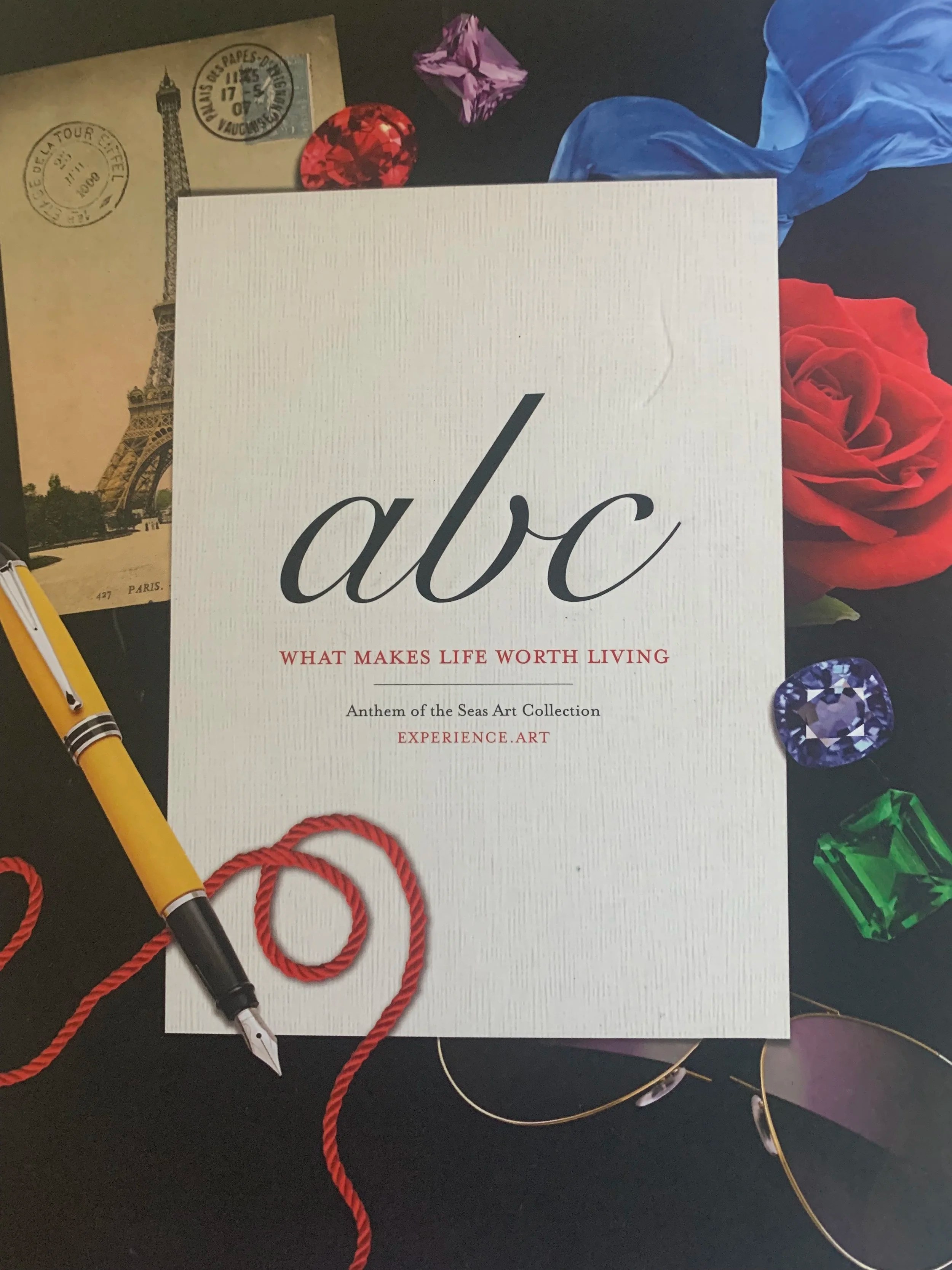 A white card with the text 'abc' in large black cursive font and 'What makes life worth living' in smaller red capital letters. Surrounding the card are various objects, including a Paris-themed postcard, a yellow fountain pen, a red string, a red rose, a purple gemstone, a green gemstone, a pair of sunglasses, a blue fabric, and a purple gemstone.