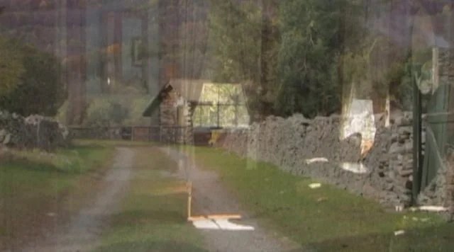 A blurry photograph showing a dirt pathway with a stone wall on the right and trees in the background.