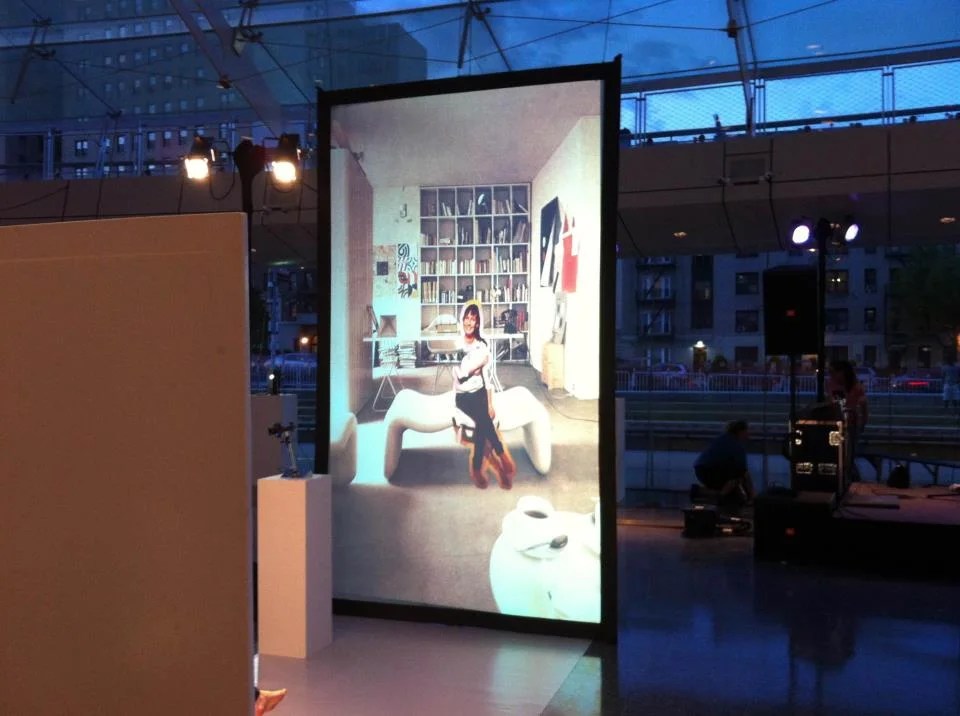 A large digital screen in an outdoor urban setting displaying a photograph of a woman standing in a modern, artistically decorated room with a white sculpture. The environment appears to be a temporary exhibition or event space at dusk.