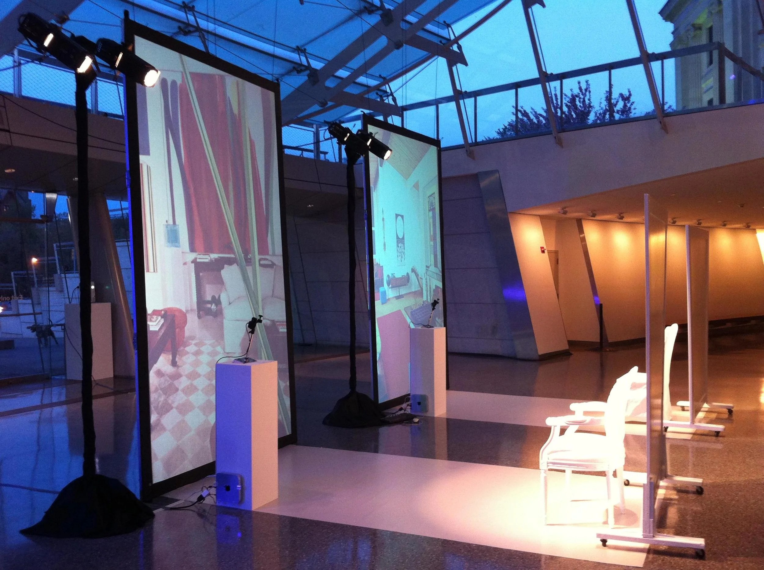 A modern indoor exhibition space with two large vertical screens displaying digital artwork or video projections, chairs, and lighting equipment, with a glass ceiling and cityscape visible outside.