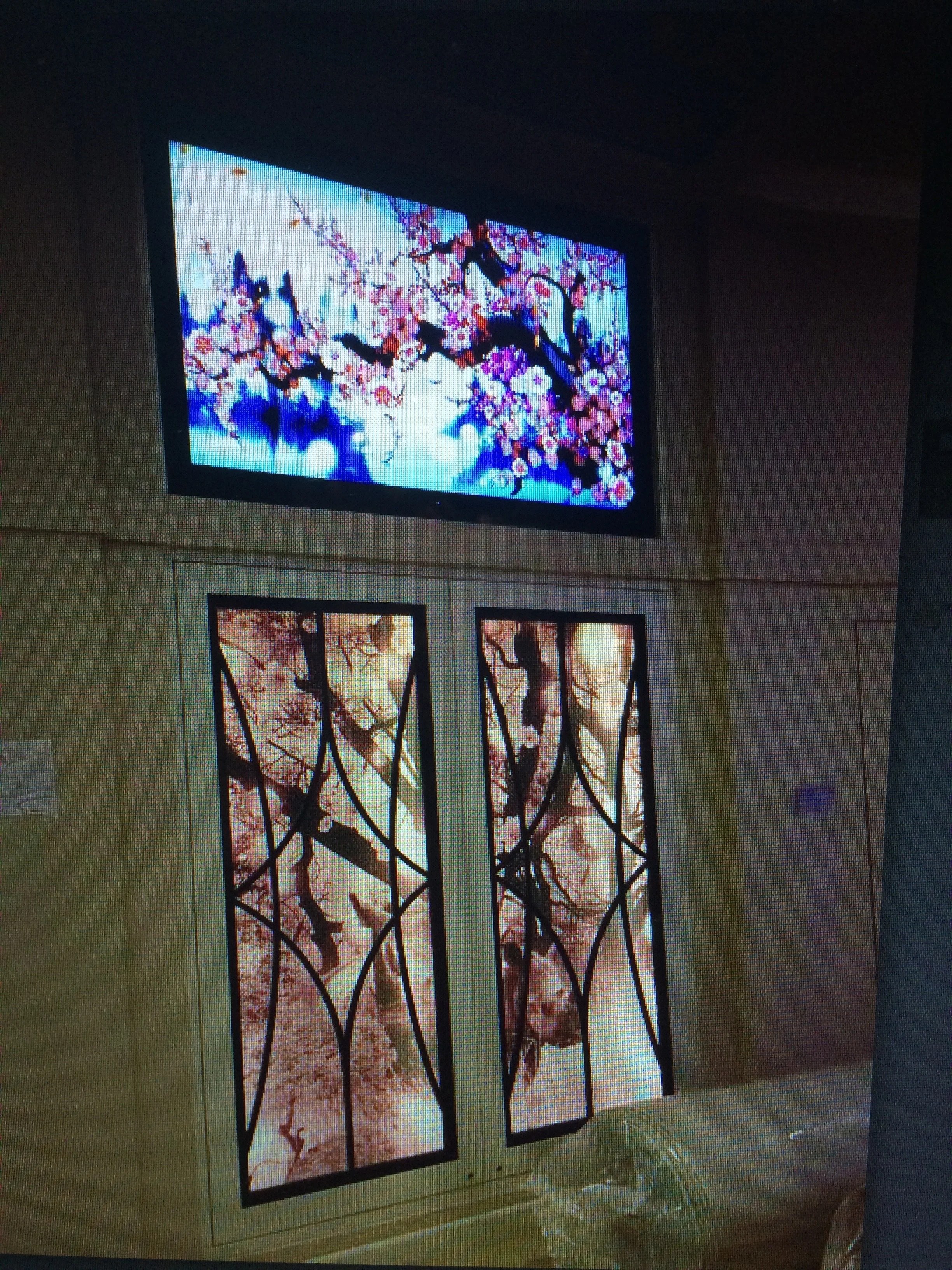Interior view of a room showing a television mounted on the wall displaying a painting of cherry blossom branches and pink flowers, and a decorative window with cherry blossom images.