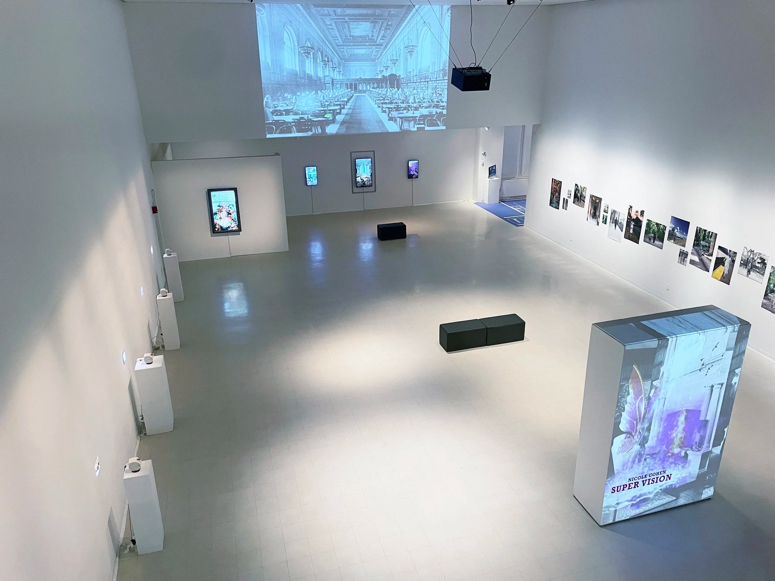 Empty art gallery with photographs and digital screens on white walls, a large projected image on the wall depicting a grand hall with benches, and a prominent display featuring Nicole Cohen's 'Super Vision' in the foreground.