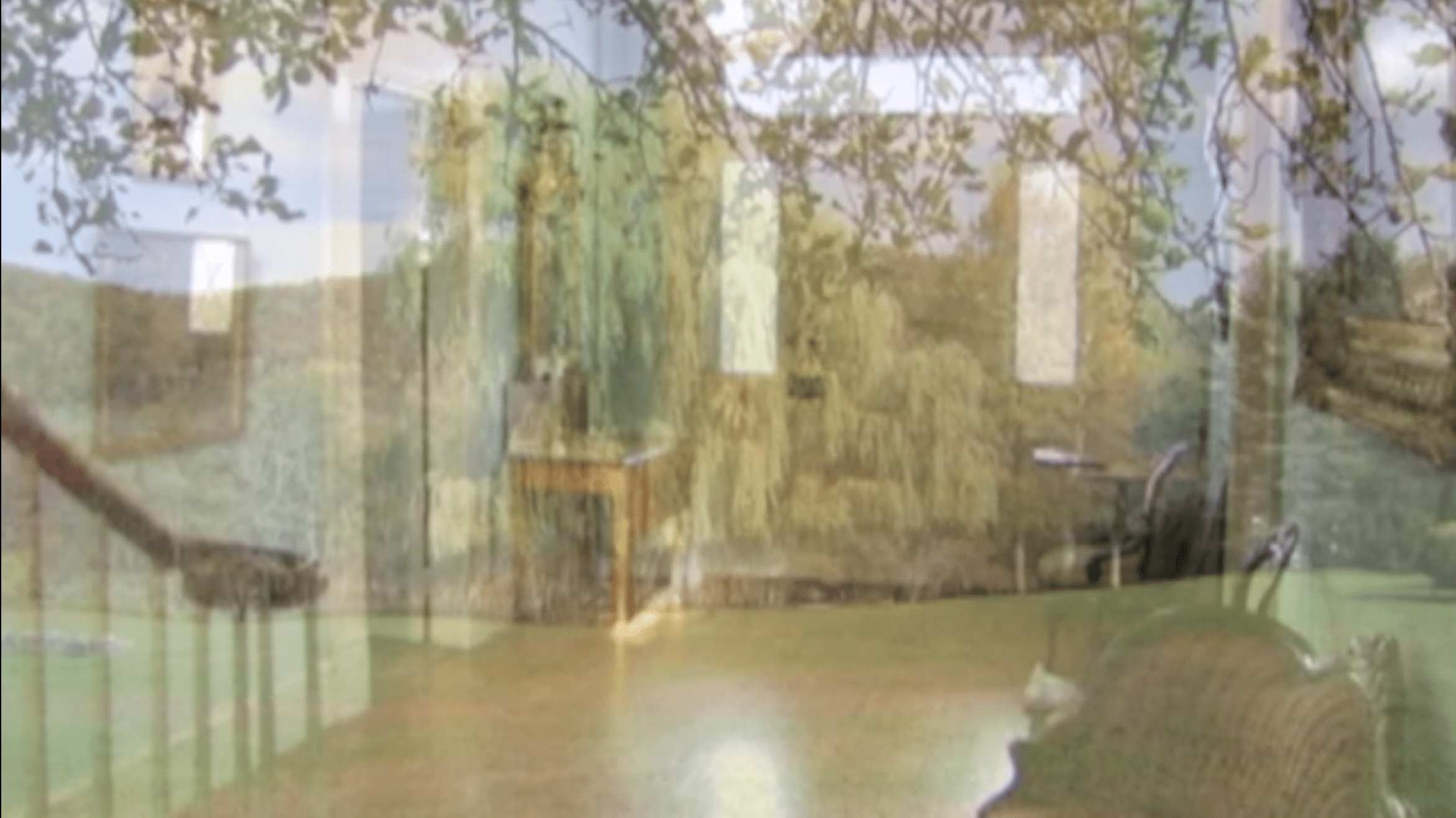 Reflected view of a room with large windows showing an outdoor landscape with trees and greenery; indoor furniture includes a table and chairs.
