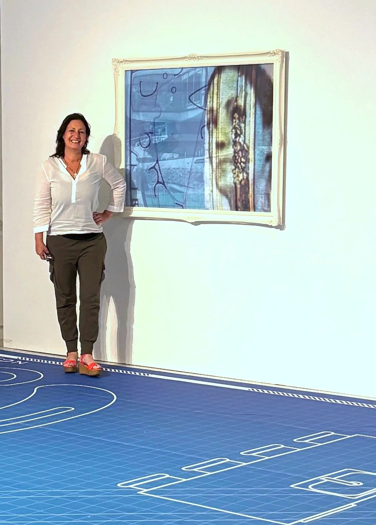 Woman standing next to a framed digital art piece on a white wall, with a blue grid-like floor in an art gallery or museum.