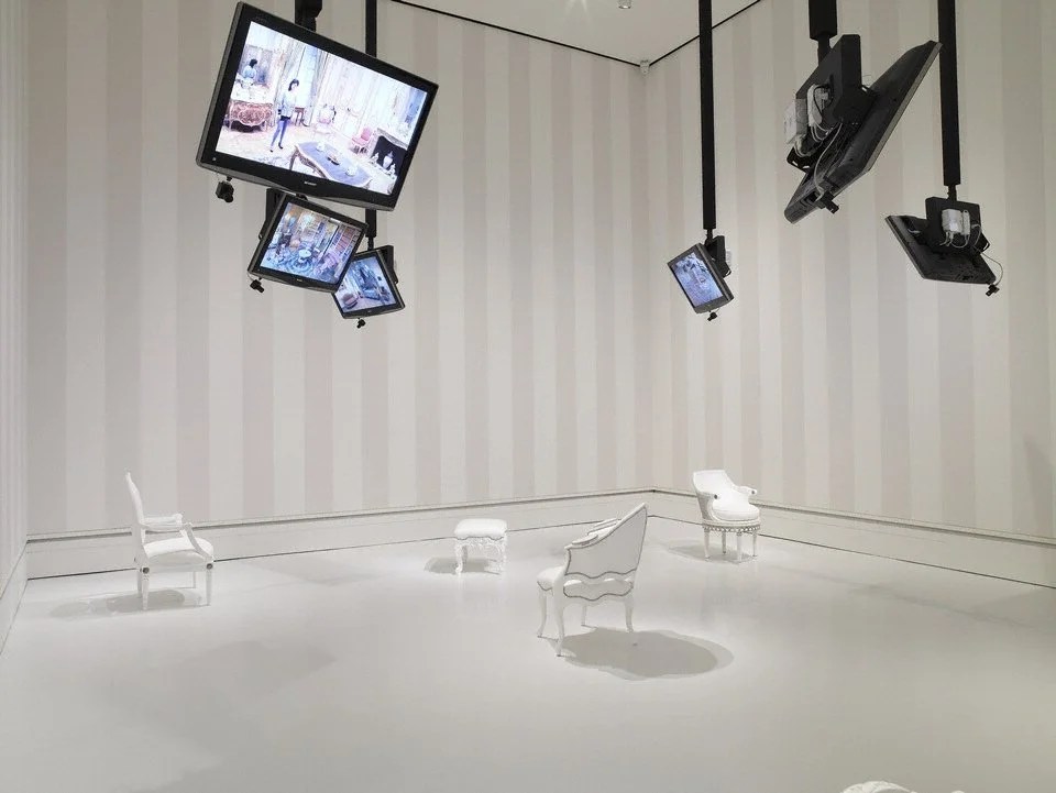 An art installation with white furniture, including a chair, a small stool, and a small chair, in a white room with striped walls. Multiple monitors and cameras are suspended from the ceiling, displaying images.