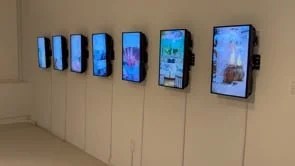 Line of seven digital screens mounted on a beige wall displaying various colorful backgrounds and images